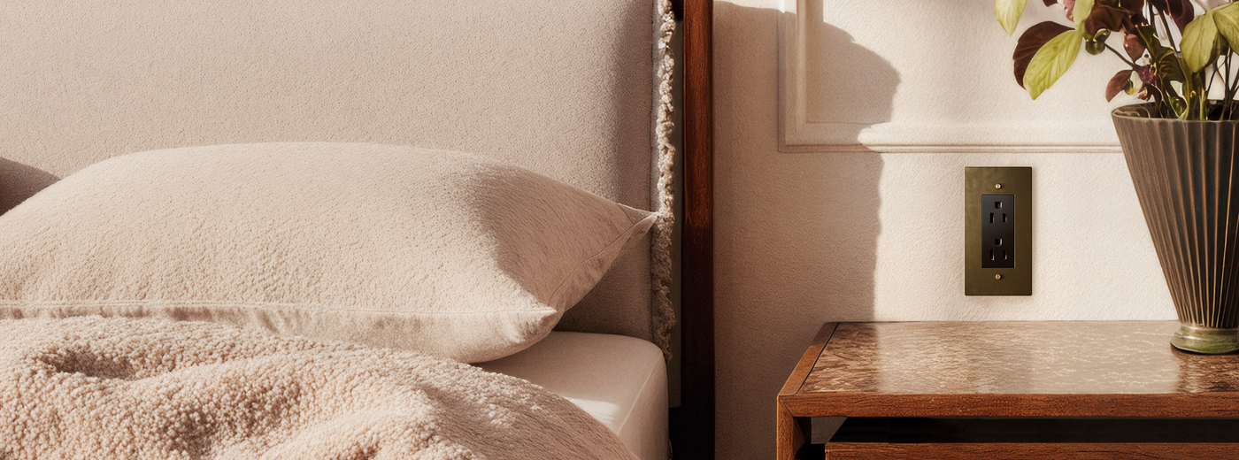 Neatly made bed with pillows and a blanket next to a wooden nightstand with a plant featuring a brass electrical outlet.