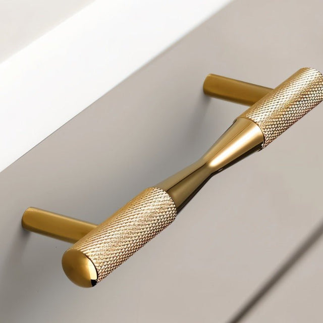 "Close-up of a brass pull bar handle with knurled detailing on a gray cabinet, showcasing its cylindrical design and tapered ends."