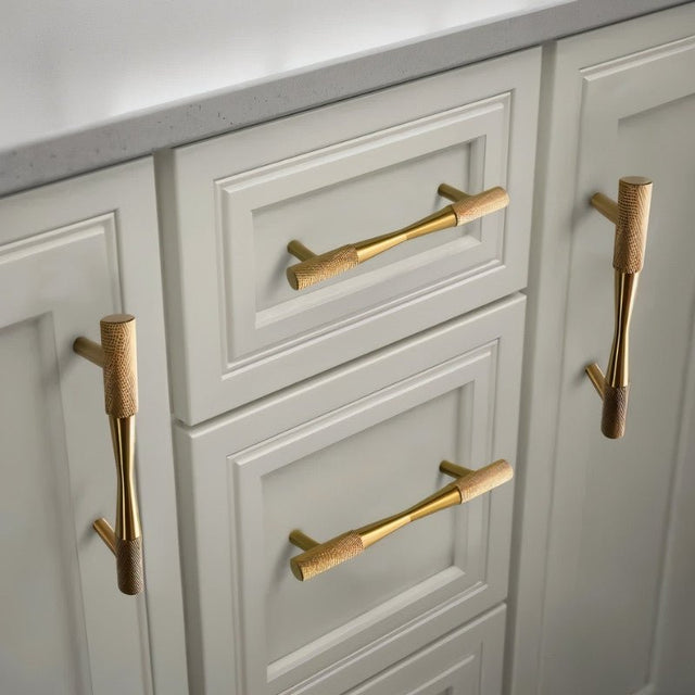 "Brass pull bar handles with knurled detailing on light gray kitchen cabinets, showcasing their cylindrical design and modern look."