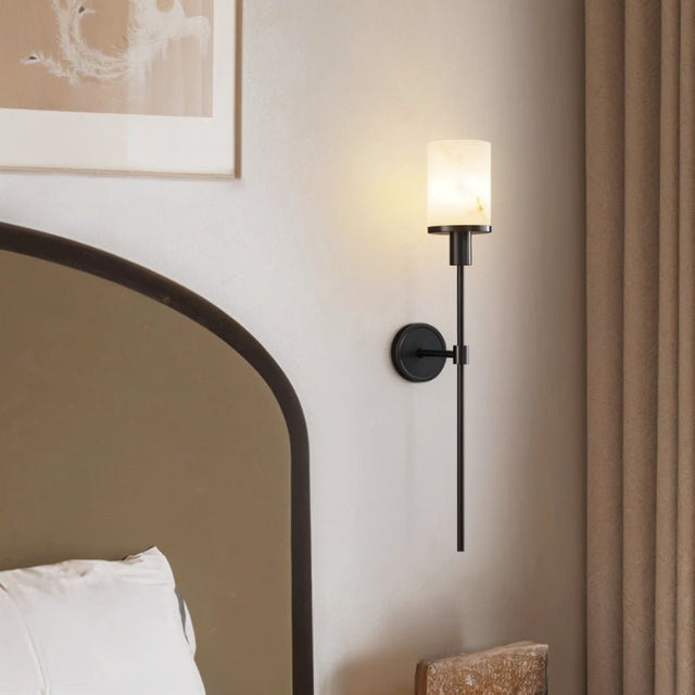 "A modern wall lamp with a cylindrical alabaster shade, mounted on a light-colored wall next to a bed with a curved headboard."