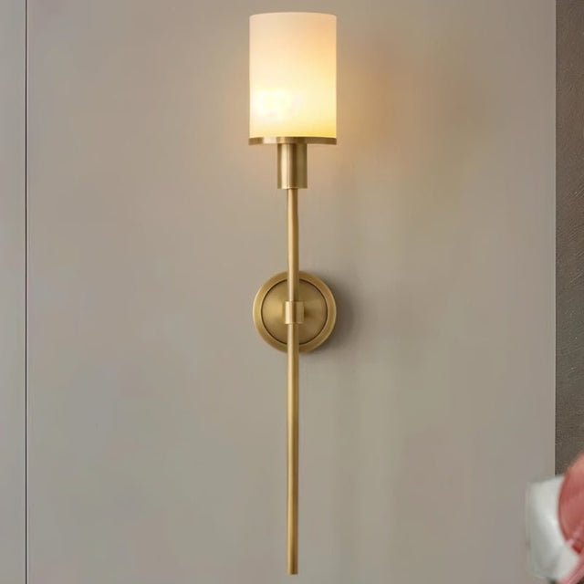 "A tall, elegant brass wall lamp with a cylindrical alabaster shade casting a warm glow on a neutral wall."