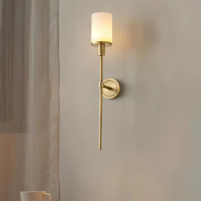 "A minimalist brass wall lamp with a cylindrical alabaster shade casting a warm, ambient glow in a modern living room."