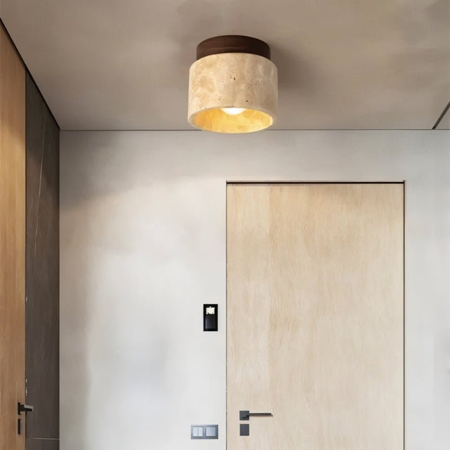 "A modern cylindrical ceiling light fixture made of yellow travertine with a dark wood top, mounted on a hallway ceiling, providing a warm, ambient glow near a closed door."