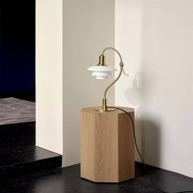 "Alya Table Lamp with a curved metallic arm, frosted glass shades, and a brass finish, placed on a wooden pedestal in a minimalist setting, creating soft, diffused lighting."