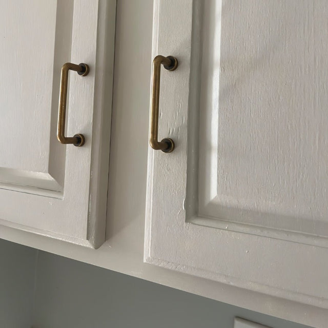 "Two minimalist antique brass pull bar handles made of durable zinc, installed on white cabinets, showcasing a slim, cylindrical design with rounded ends."
