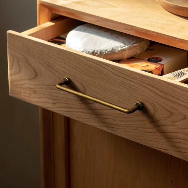 "A minimalist antique brass pull bar handle made of durable zinc, installed on a light wood drawer with an open drawer showcasing items inside."
