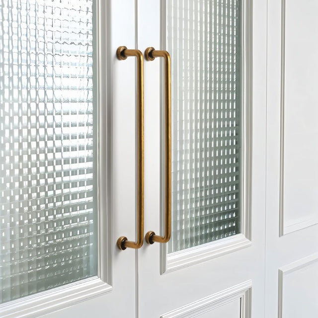 "Two long, minimalist antique brass pull bar handles made of durable zinc, installed on white framed glass doors, featuring a slim, cylindrical design with rounded ends."