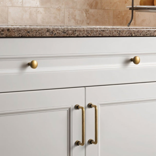 "Minimalist antique brass pull bar handles and knobs made of durable zinc, installed on white kitchen cabinets with a granite countertop."