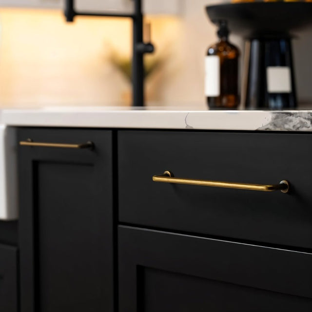 "Two minimalist antique brass pull bar handles made of durable zinc, installed on black kitchen cabinets with a white countertop and a blurred background featuring a black faucet and a fruit bowl."