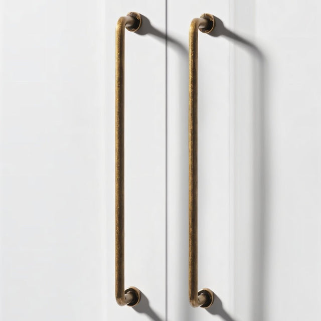 "Two long, minimalist antique brass pull bar handles made of durable zinc, installed on white doors, featuring a slim, cylindrical design with rounded ends."