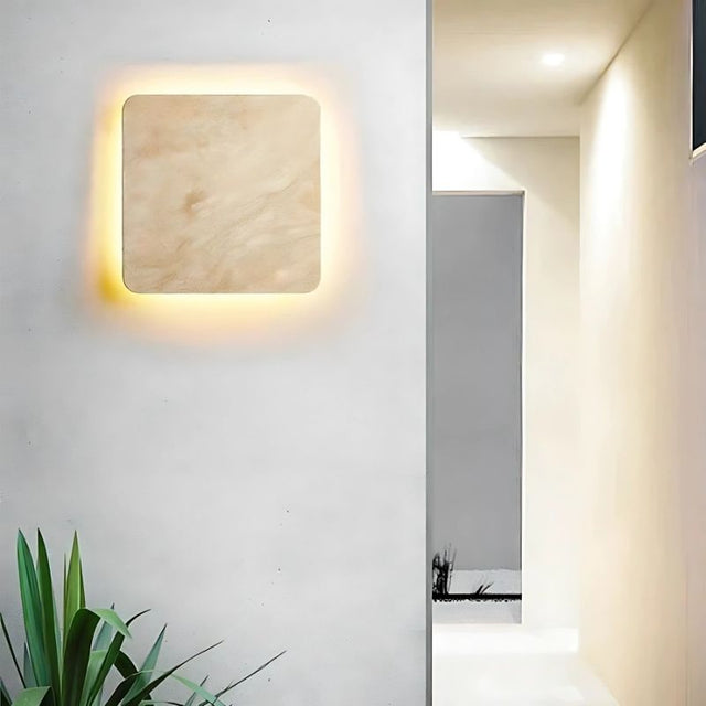 "A square Amon travertine wall lamp with a smooth, minimalist design illuminates a modern hallway with white walls, casting a warm glow near a green potted plant and a glimpse into another room."