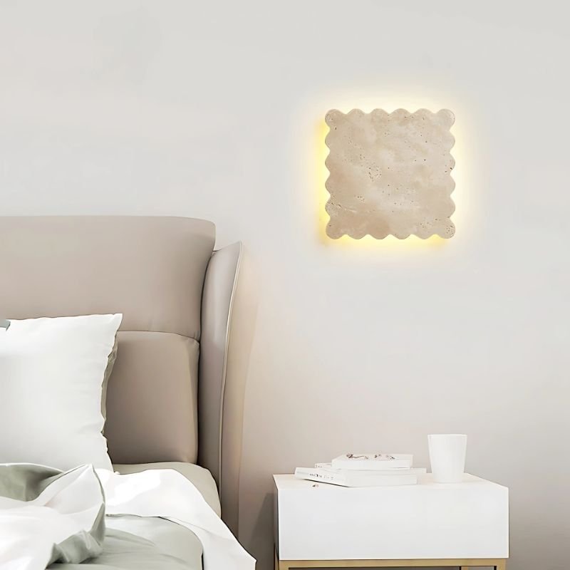 "The Amon travertine wall lamp, showcasing a unique square shape with scalloped edges, illuminates a modern bedroom with a neutral-toned bed and a white nightstand featuring books and a white cup."