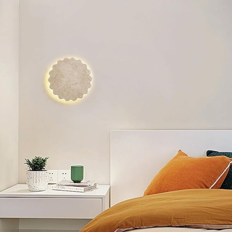 "The Amon travertine wall lamp, with its scalloped, flower-like design, complements a modern bedroom featuring a white nightstand with a potted plant, books, and a green container, alongside an orange and brown bed ensemble."