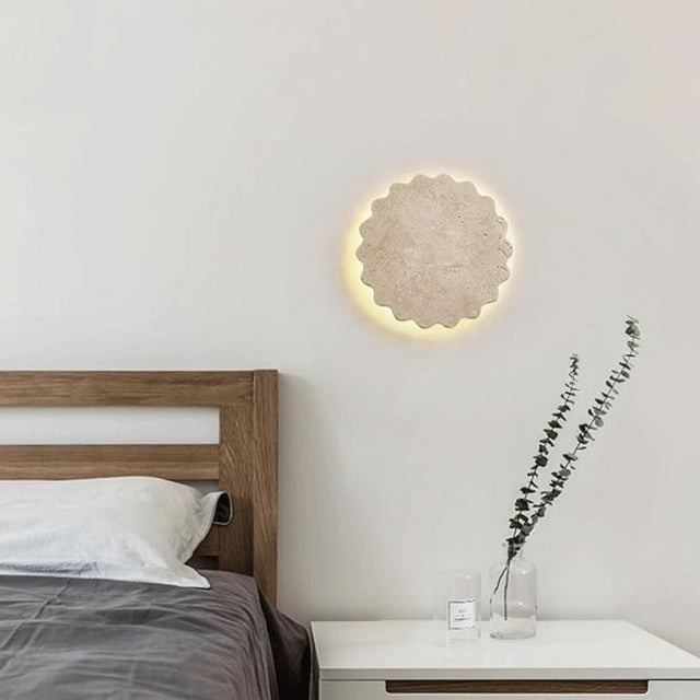"The Amon travertine wall lamp, with its scalloped, flower-like design, illuminates a modern bedroom nightstand with a glass vase and dried floral arrangement, creating a warm and inviting atmosphere."