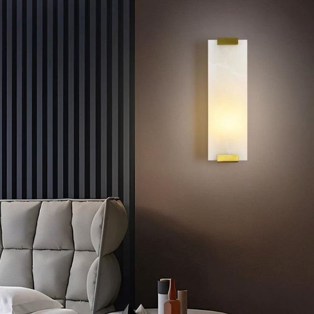 "A modern wall lamp with a rectangular alabaster panel and brass accents, providing a soft, ambient glow in a contemporary bedroom."