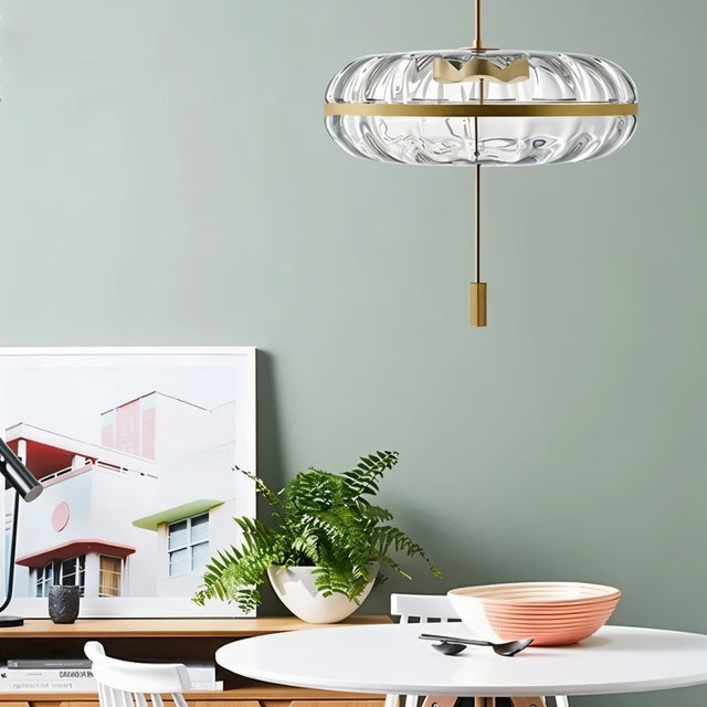 "Sculptural glass pendant light with rippled texture and metal frame, hanging above a dining table with plants and artwork."