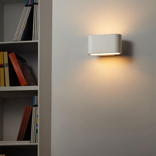 "White gypsum Arcat wall lamp with a rounded rectangular shape, emitting soft light on a wall near a bookshelf."