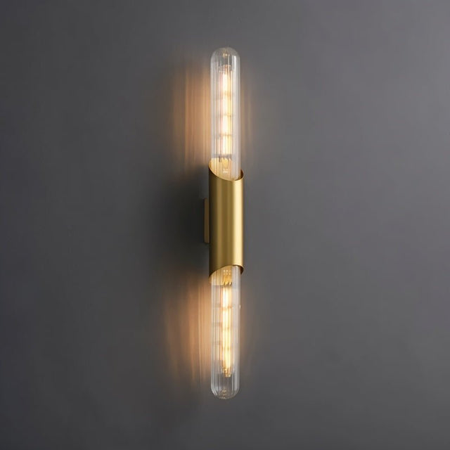 "A modern brass wall lamp with a vertical ribbed glass tube on a dark gray wall."