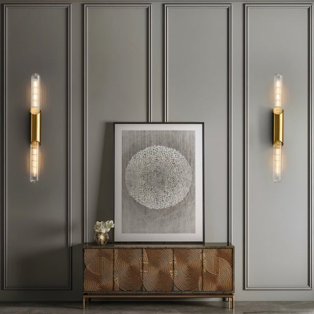 "Two modern brass wall lamps with vertical glass tubes flanking a framed artwork in a hallway."