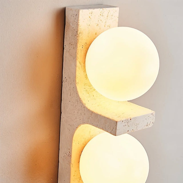"Close-up of a modern travertine wall lamp with two spherical white glass shades, showcasing the texture of the natural stone and the soft glow of the light."