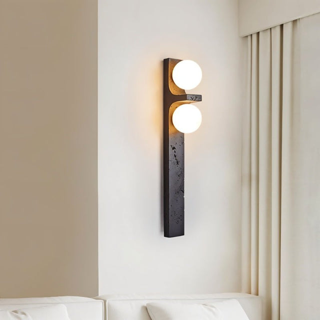 "A modern travertine wall lamp with two spherical white glass shades mounted on a vertical travertine base, illuminating a bedroom with soft, ambient light."