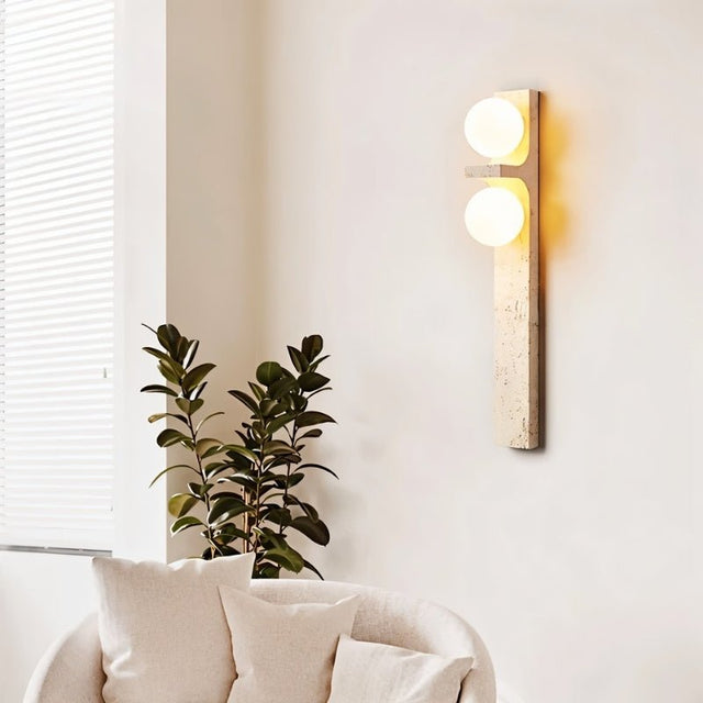 "A modern travertine wall lamp with two spherical white glass shades mounted on a vertical travertine base, illuminating a modern living room."