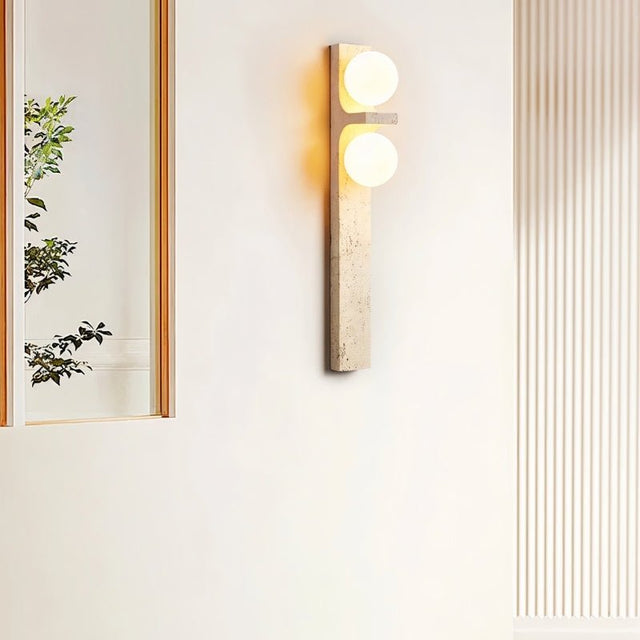 "A modern travertine wall sconce with two spherical white glass shades mounted on a vertical travertine base."