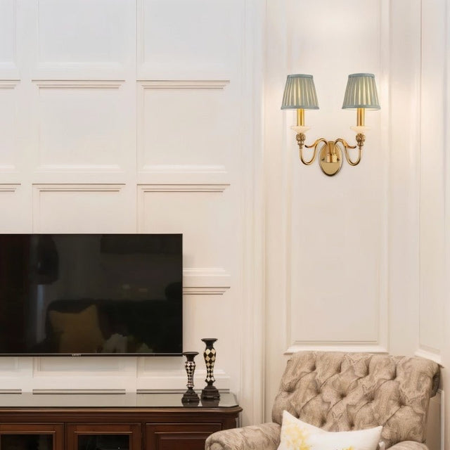 "A double-arm brass wall lamp with jade accents and fabric shades, mounted on a wall in a living room with a television."