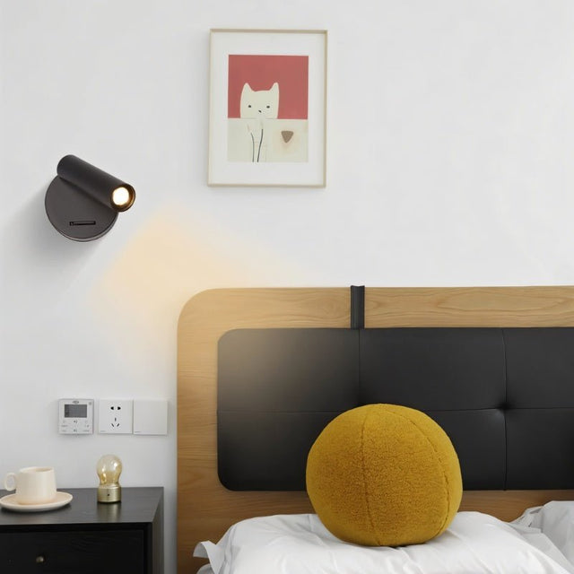 "A black aluminum wall lamp with an adjustable cylindrical head, installed above a bedside table with a cup and a light bulb, next to a bed with a yellow round pillow and a framed cat picture."