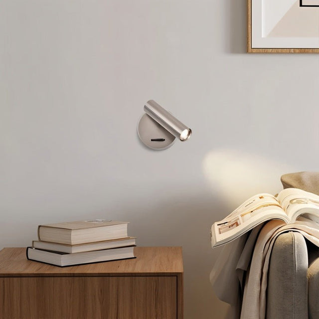 "A nickel aluminum wall lamp with an adjustable cylindrical head, installed above a bedside table with books and a magazine, next to a gray couch."