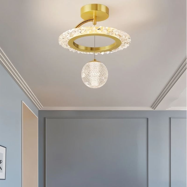 "A gold ceiling light with a circular frame and a hanging crystal sphere, installed on a ceiling in a room with gray walls."