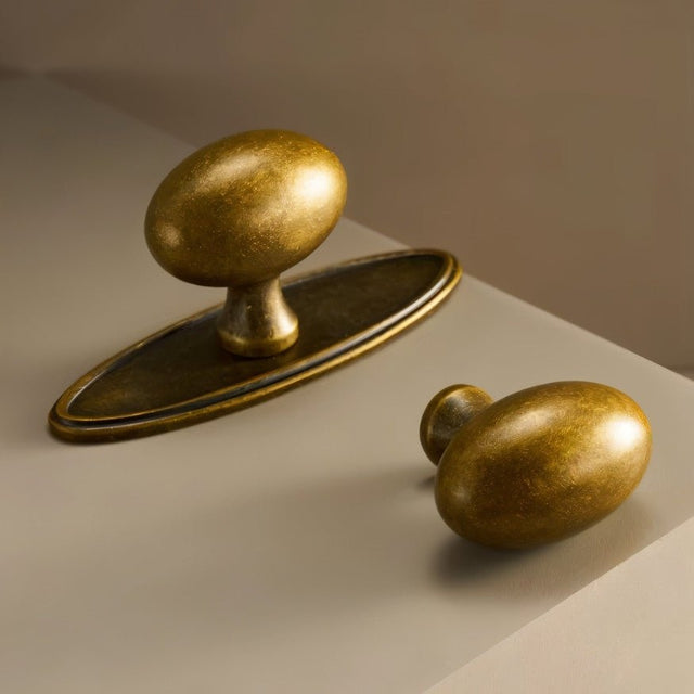 "Close-up of a pair of oval antique brass knobs, one with a backplate, showcasing their smooth, ergonomic design for cabinets and drawers."