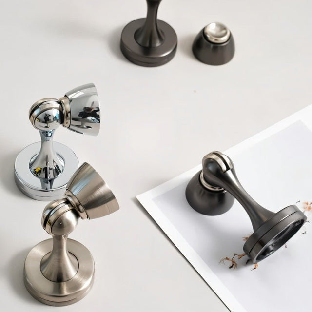 "Several sleek, zinc-alloy magnetic door stops with contoured designs in various finishes, lying on a white surface, designed to prevent door slams and wall damage in modern interiors."