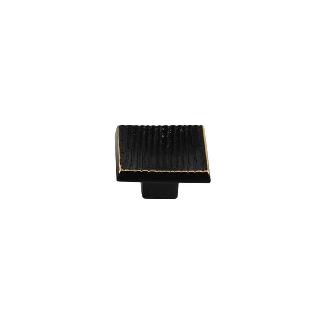 "Studio shot of a single black square cabinet knob with a ridged, textured surface and brass edges, on a white background."