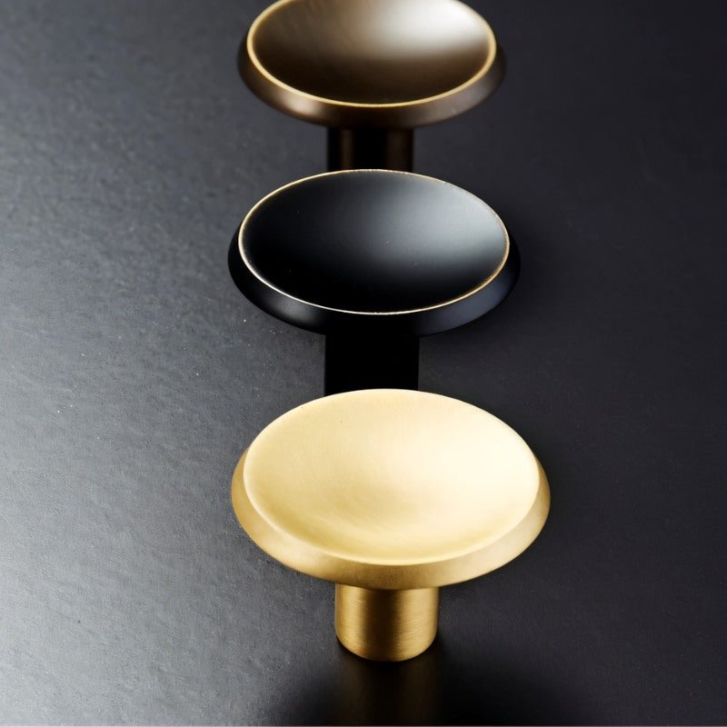 "Top view of three circular brass knobs in bronze, black, and gold with concave surface and chamfered edges."