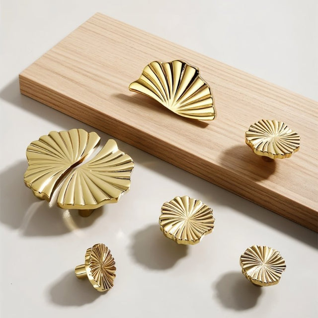 "A variety of gold brass fan-shaped drawer pulls arranged on a light surface, showcasing different sizes and styles."