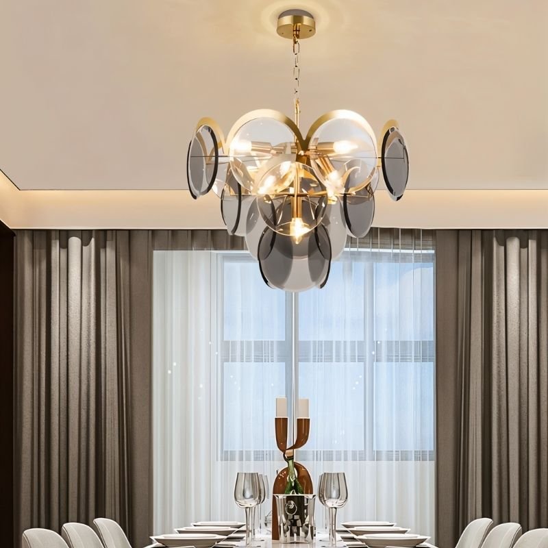 "A modern chandelier with layered smoked glass discs hanging above a dining table in a contemporary dining room with sheer curtains."