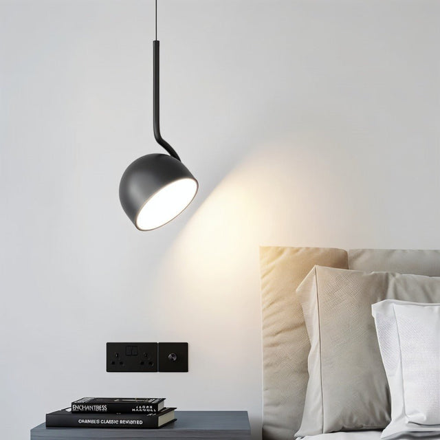 "A black, curved iron pendant light with a dome-shaped shade illuminates a bedside reading nook with books stacked on a small table."