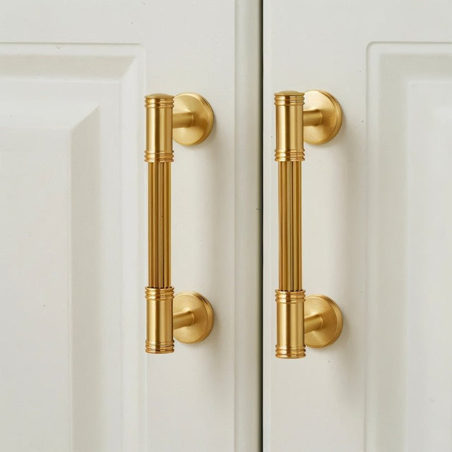 "Two brass pull bars with grooved detailing mounted on a white cabinet door, showcasing a sophisticated and timeless design."