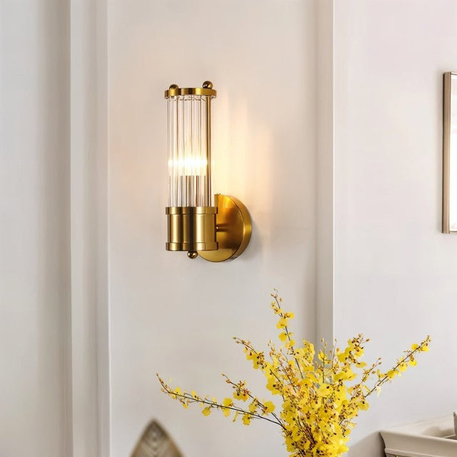 "A modern gold wall lamp with clear glass panels, mounted on a white wall with a yellow flower arrangement below."