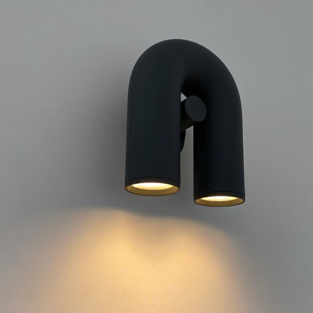 "A close-up of a black, contemporary wall lamp with a looped design, showing the dual light sources illuminated against a gray background."