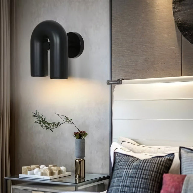 "A black, contemporary wall lamp with a looped design, mounted on a textured gray wall in a modern bedroom setting."