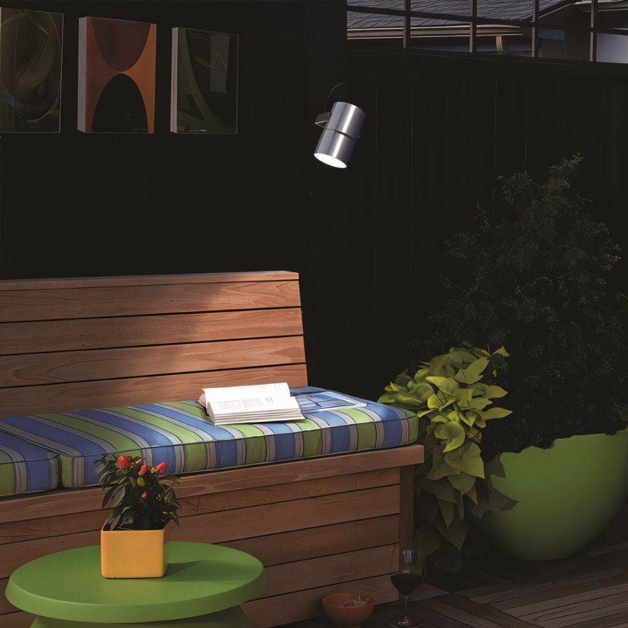 "Silver outdoor spotlight mounted on a dark wall, illuminating a patio with a wooden bench, plants, and a green table."