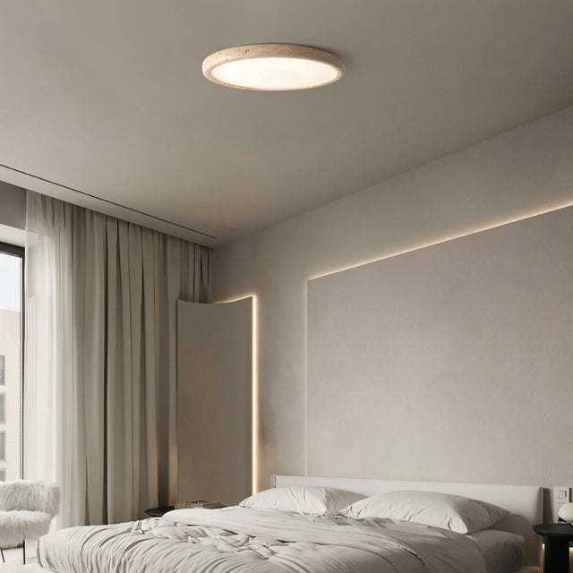 "A modern travertine flush mount ceiling light with a circular design, providing soft ambient lighting in a minimalist bedroom with a large window and white bedding."
