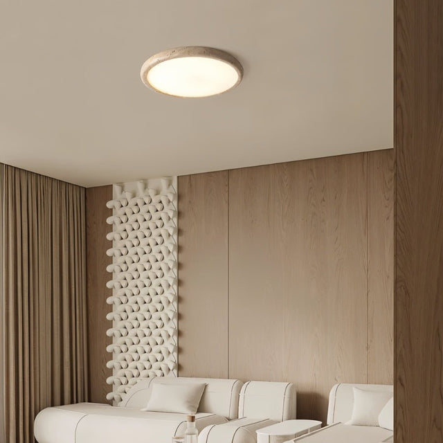"A modern travertine flush mount ceiling light with a circular design, providing ambient lighting in a contemporary living room with a textured wall and a white sofa."