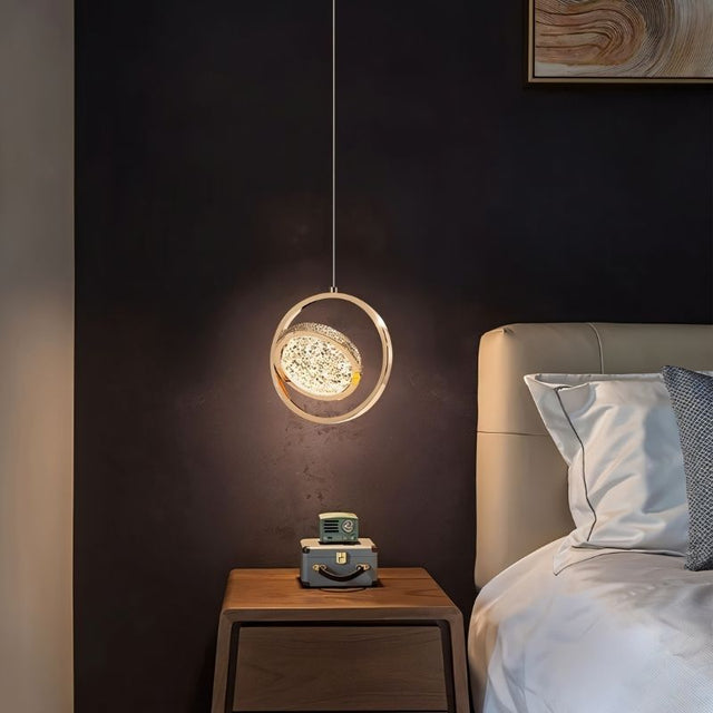 "A gold ring pendant light with a crystal-like center, hanging above a nightstand in a bedroom with a dark wall."