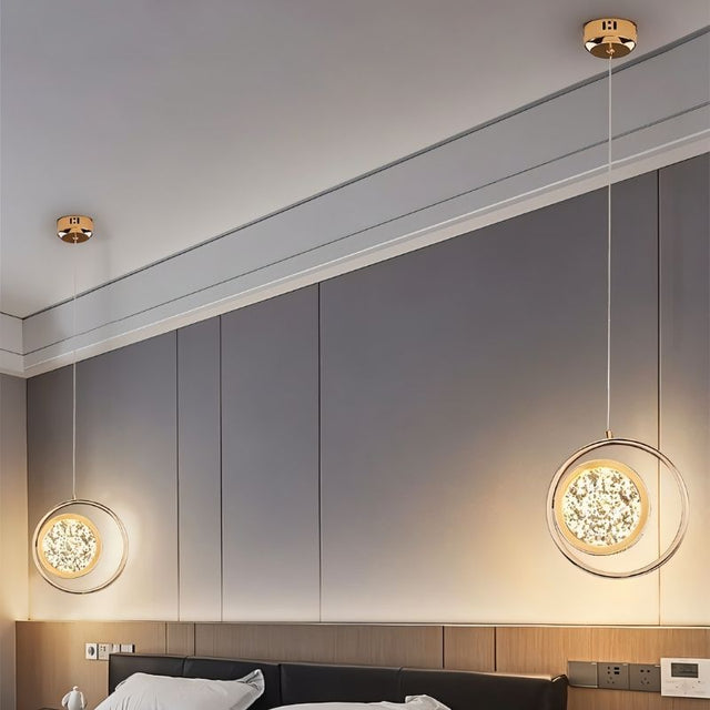 "Two gold ring pendant lights with crystal-like centers, hanging on either side of a bed in a room with a gray paneled wall."