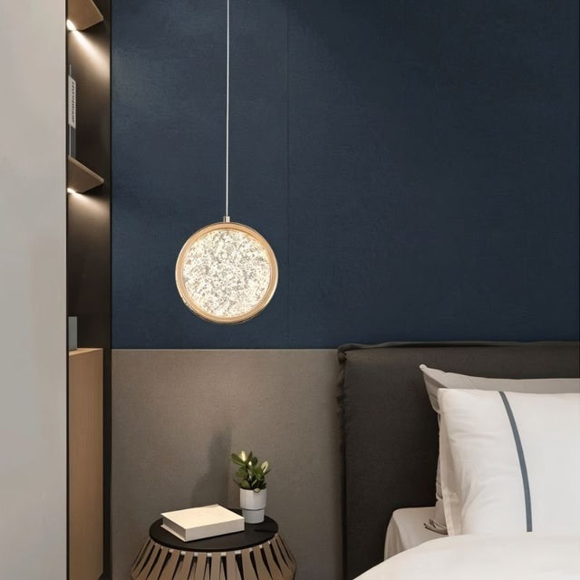 "A gold ring pendant light with a crystal-like center, hanging next to a bed in a room with a dark blue wall."