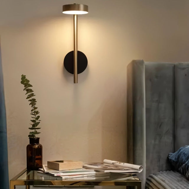 "A gold and black wall lamp with a round base and a vertical light source, installed above a nightstand with a plant, magazines, and a box, next to a gray tufted headboard in a bedroom."