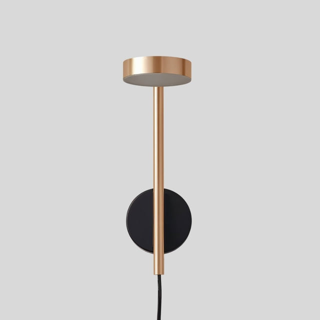 "A gold and black wall lamp with a round base and a vertical light source, shown against a gray background."